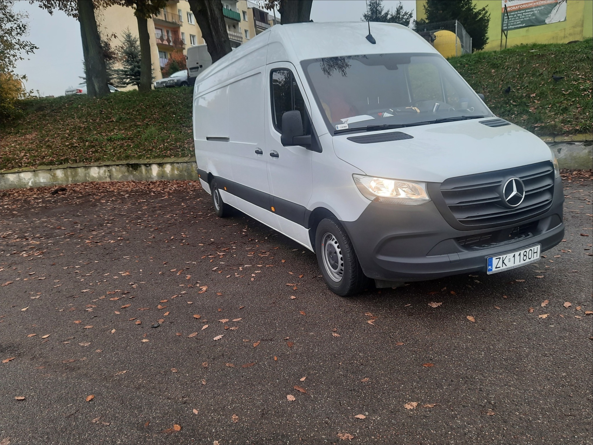 Biały samochód dostawczy Mercedes-Benz Sprinter zaparkowany na parkingu pokrytym opadłymi liśćmi, z widoczną tablicą rejestracyjną ZK 1180H i budynkiem w tle.