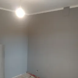 Świeżo pomalowane na szaro ściany w rogu pokoju, widoczna rolka folii malarskiej, gniazdko elektryczne i listwy przypodłogowe.