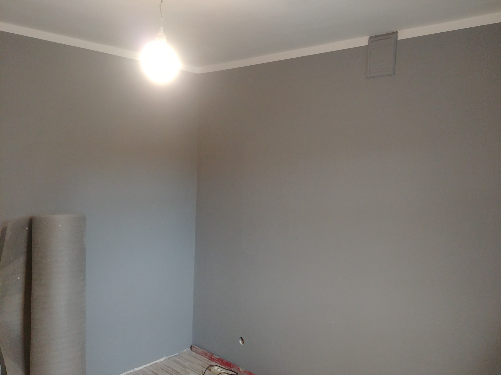 Świeżo pomalowane na szaro ściany w rogu pokoju, widoczna rolka folii malarskiej, gniazdko elektryczne i listwy przypodłogowe.