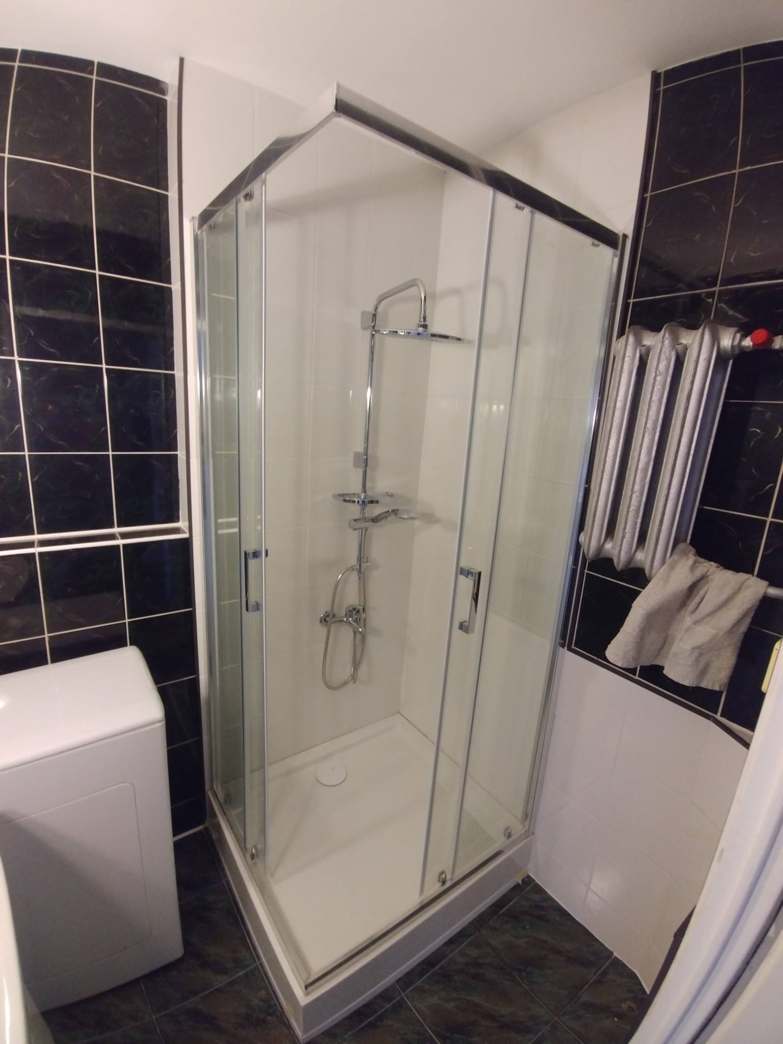 Narożna kabina prysznicowa z chromowanymi profilami i deszczownicą, widoczna pralka obok, ciemne płytki na ścianach, grzejnik żeberkowy z powieszonym ręcznikiem.