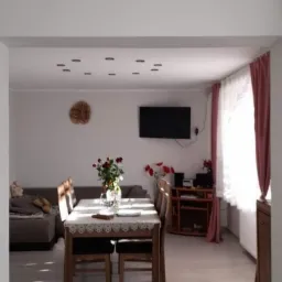 Jasne wnętrze pokoju dziennego z drewnianym stołem nakrytym koronkowym obrusem, szarym narożnikiem i telewizorem na ścianie, oświetlenie punktowe w suficie.