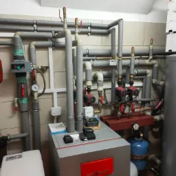Węzeł cieplny z piecem Viessmann, licznymi rurami z izolacją, zaworami, pompami obiegowymi, manometrami, zbiornikiem wyrównawczym oraz narzędziami na obudowie pieca.