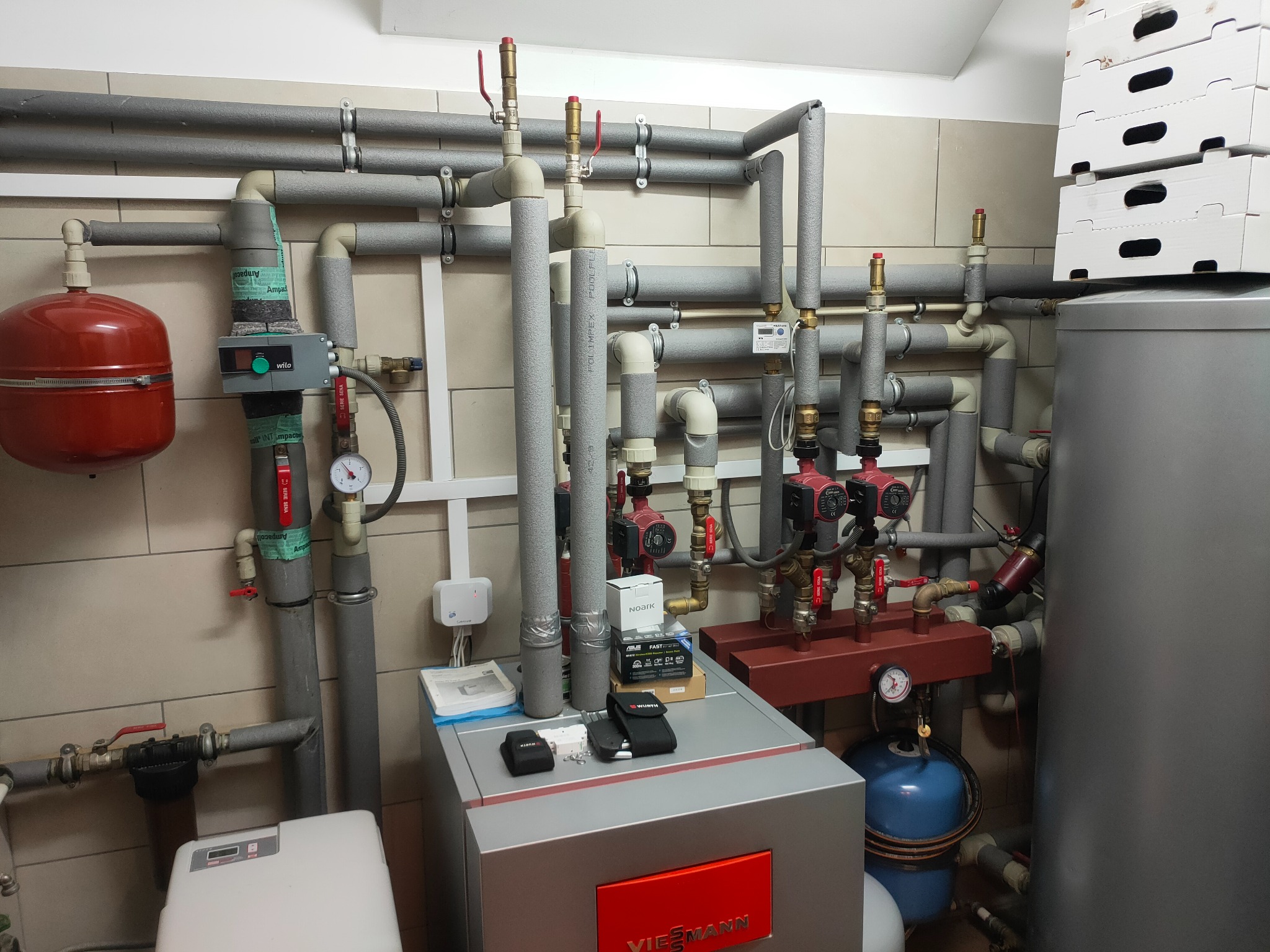 Węzeł cieplny z piecem Viessmann, licznymi rurami z izolacją, zaworami, pompami obiegowymi, manometrami, zbiornikiem wyrównawczym oraz narzędziami na obudowie pieca.