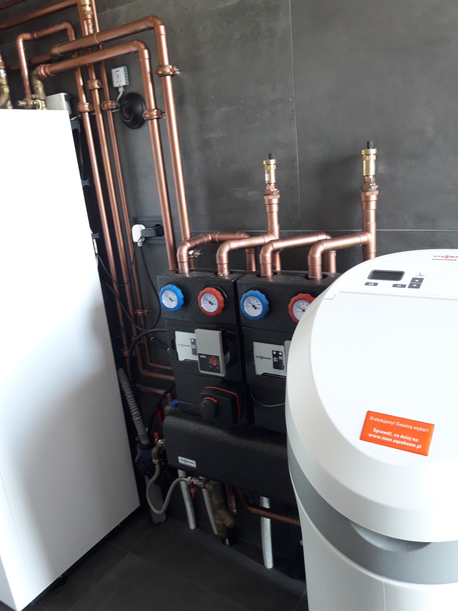 Nowoczesna kotłownia z miedzianymi rurami i urządzeniem uzdatniającym wodę Viessmann Aquahome w Poznaniu, widoczne manometry i liczniki, szara ściana w tle.