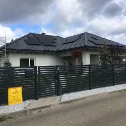 Dom jednorodzinny z ciemną dachówką i panelami słonecznymi na dachu, widoczna aluminiowa drabina oparta o dach, szara, nowoczesna brama i żółta skrzynka gazowa na chodniku.