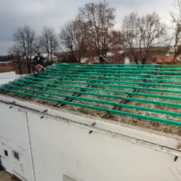 Ekipa dekarzy montuje zieloną konstrukcję nośną pod nowe pokrycie dachu na płaskim dachu budynku, widok z góry na plac budowy w pochmurny dzień.