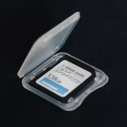 Karta pamięci 128GB w przezroczystym etui, z logo 'SOMA media YOUR STORIES', leżąca na czarnym tle. Detal produktu identyfikacji wizualnej.