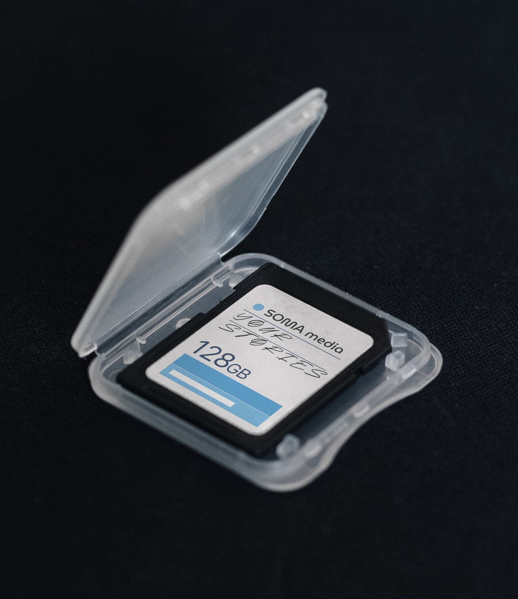 Karta pamięci 128GB w przezroczystym etui, z logo 'SOMA media YOUR STORIES', leżąca na czarnym tle. Detal produktu identyfikacji wizualnej.