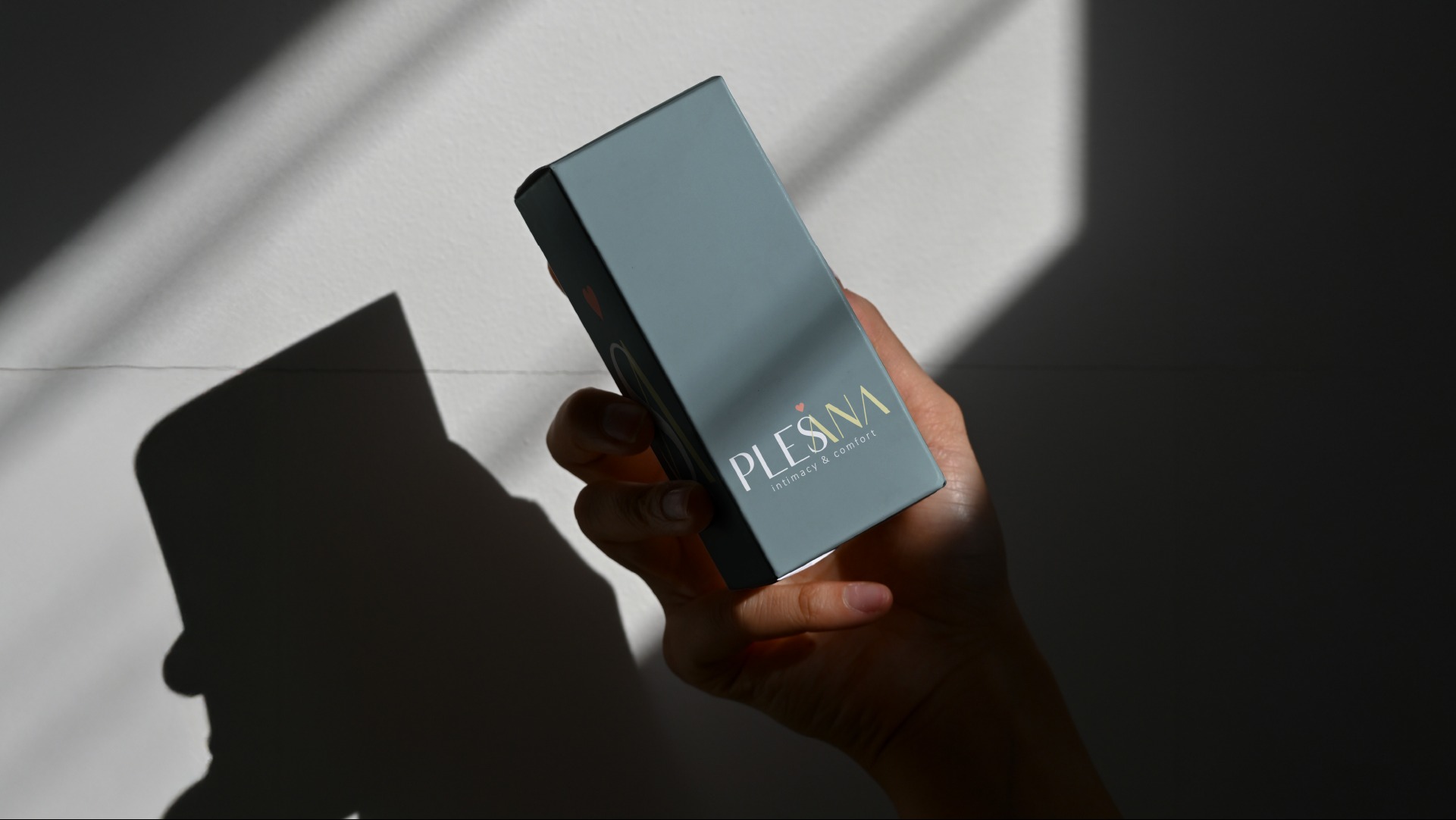 Dłoń trzyma pudełko z logo Plesana w naturalnym świetle. Minimalistyczny design, szaro-zielone tło, wyraźne cienie tworzą kontrast. Subtelna elegancja.