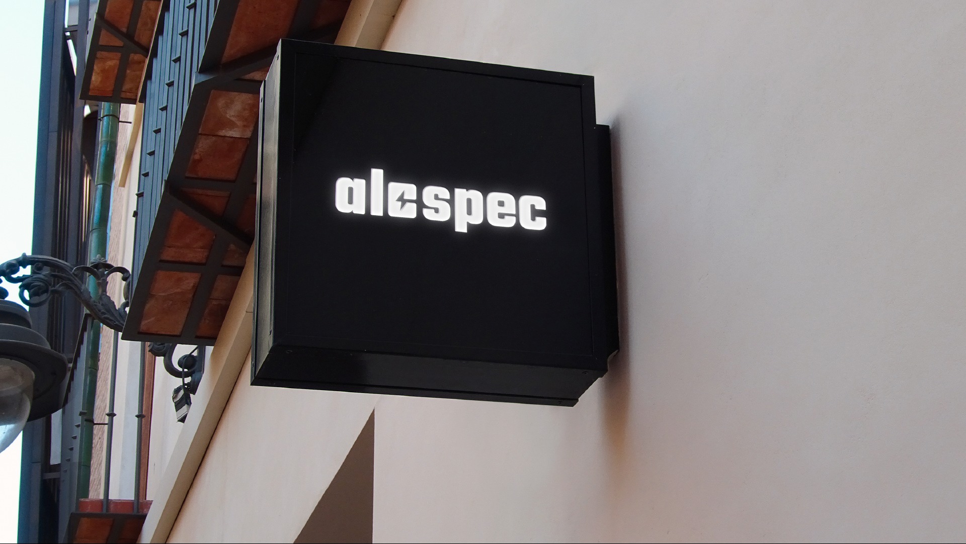 Czarny, podświetlany szyld reklamowy z nazwą 'alespec' na elewacji budynku z elementami architektonicznymi w tle. Kątowe ujęcie.