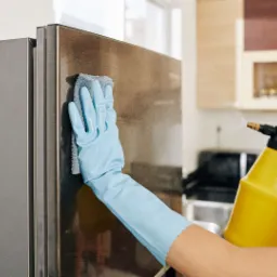 Osoba w niebieskich rękawicach czyści stalową lodówkę szarą ściereczką i żółtym spryskiwaczem w kuchni.