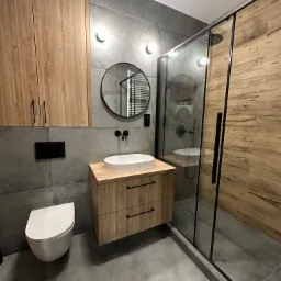 Nowoczesna łazienka z szarymi płytkami, drewnianymi szafkami i kabiną prysznicową typu walk-in z elementami drewna, okrągłe lustro nad umywalką, podwieszana toaleta.