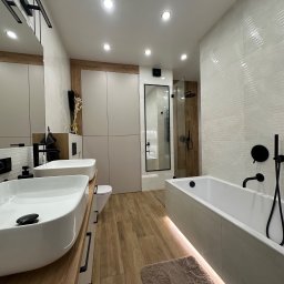 ASK BUD - Nowoczesna łazienka z drewnianymi akcentami: podwójna umywalka nablatowa, wanna z podświetleniem LED, kabina prysznicowa w czarnej ramie i płytki imitujące drewno.