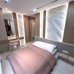 ASK BUD - Nowoczesna sypialnia z łóżkiem, toaletką i oświetleniem LED. Ściana z dekoracyjną strukturą i drewnianymi panelami. Minimalistyczny design, jasne kolory, eleganckie wykończenie.