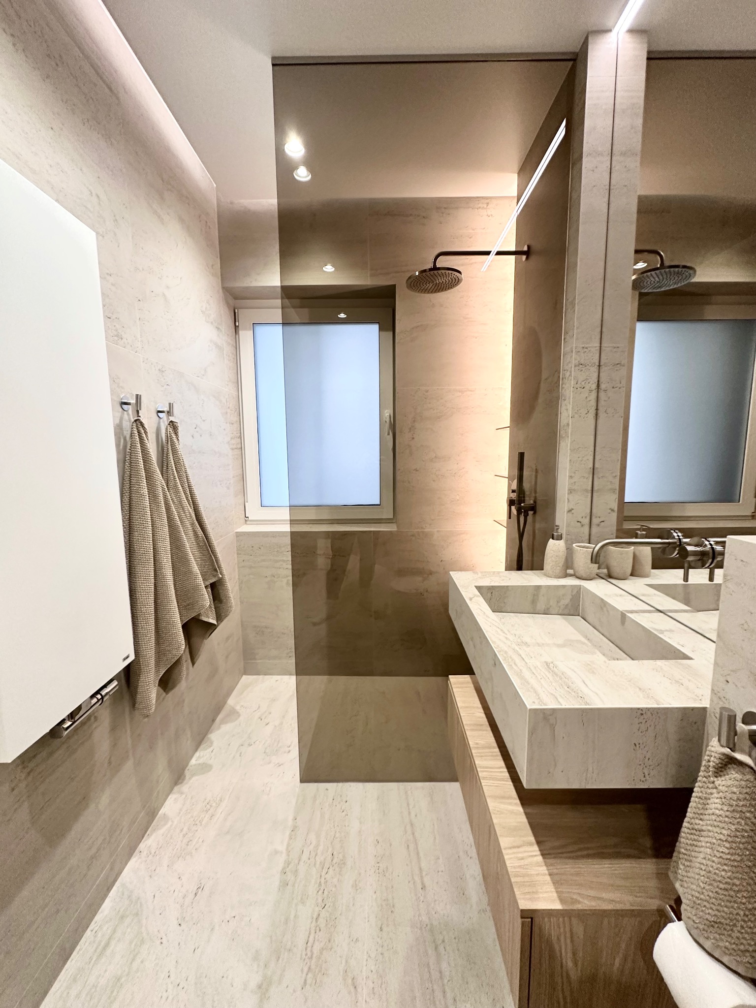 Nowoczesna łazienka z beżowymi płytkami imitującymi kamień, deszczownicą, umywalką z trawertynu i białym grzejnikiem drabinkowym.