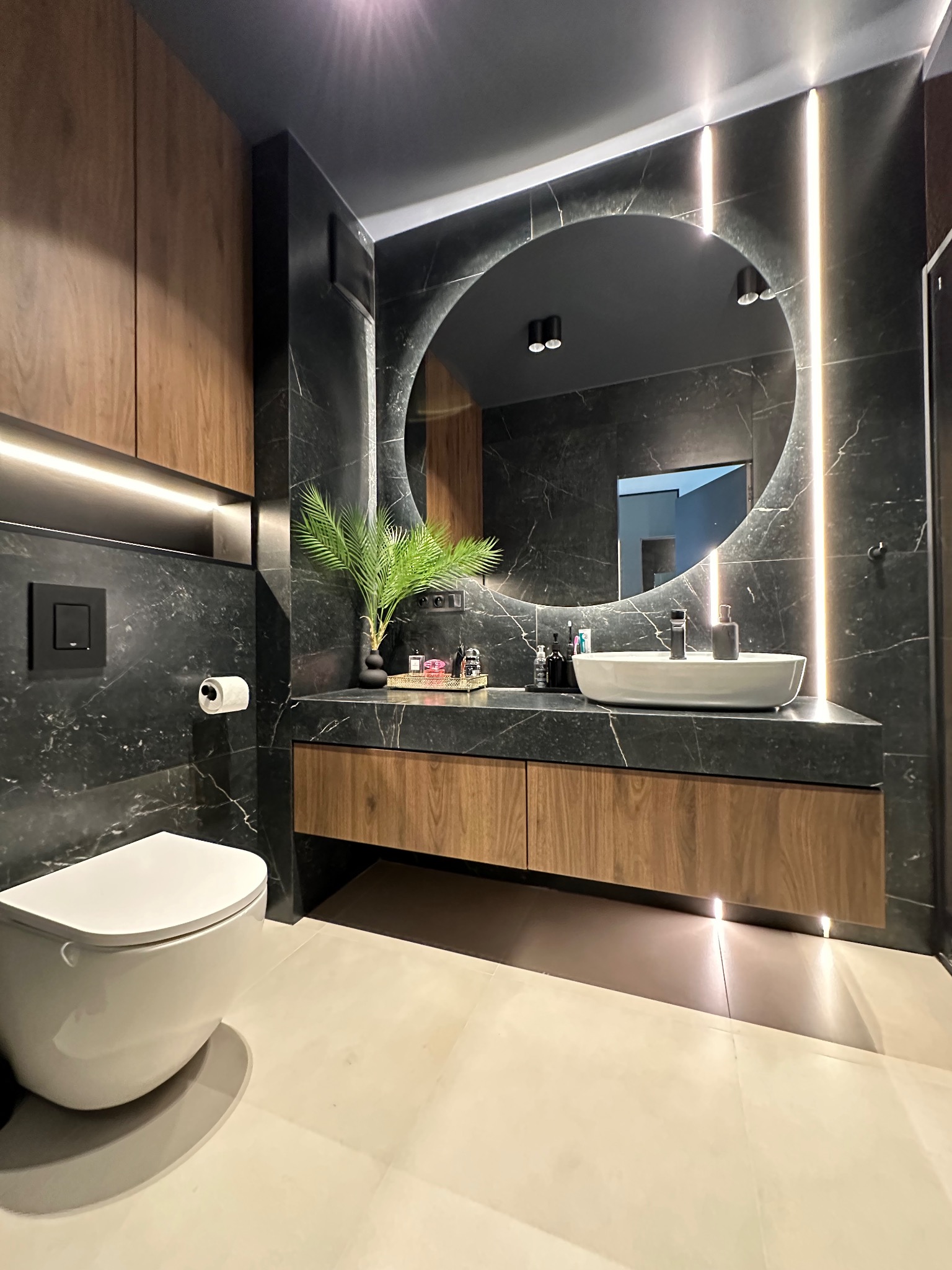 Nowoczesna łazienka z czarnymi marmurowymi ścianami, okrągłym podświetlanym lustrem, drewnianymi szafkami pod umywalką i wiszącą toaletą; akcent roślinny w postaci palmy w czarnym wazonie.