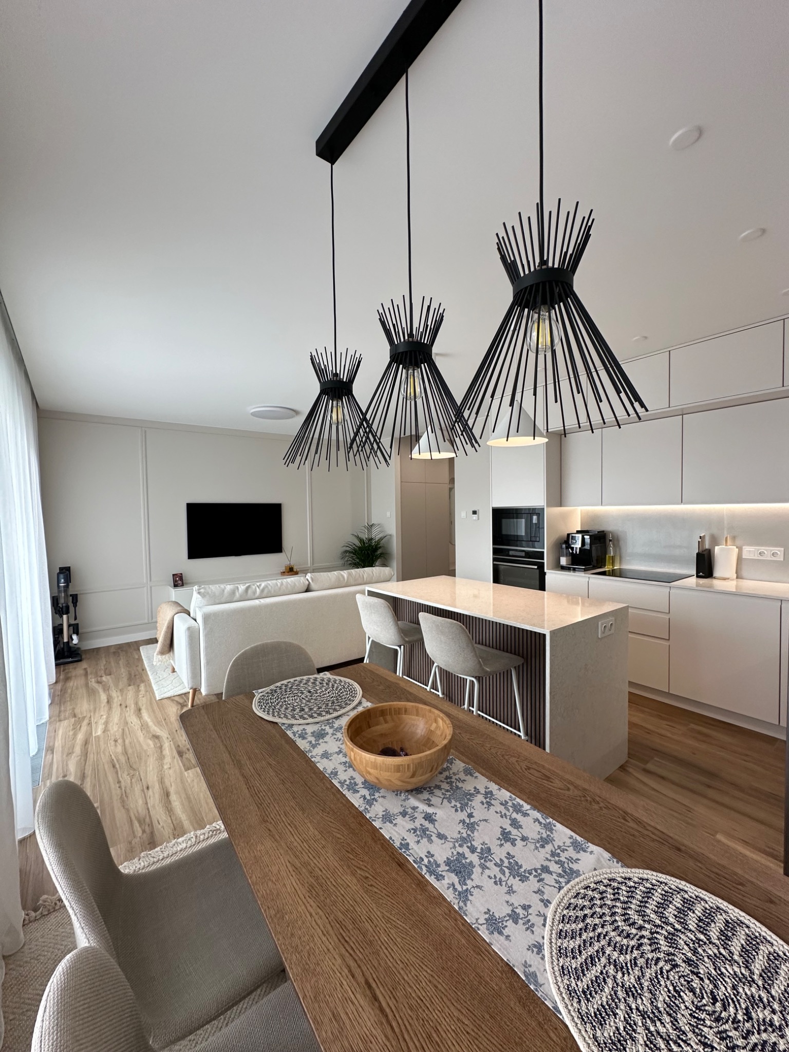 Jasne, przestronne wnętrze domu z drewnianym stołem, designerskimi lampami wiszącymi nad wyspą kuchenną i minimalistycznym salonem w tle.