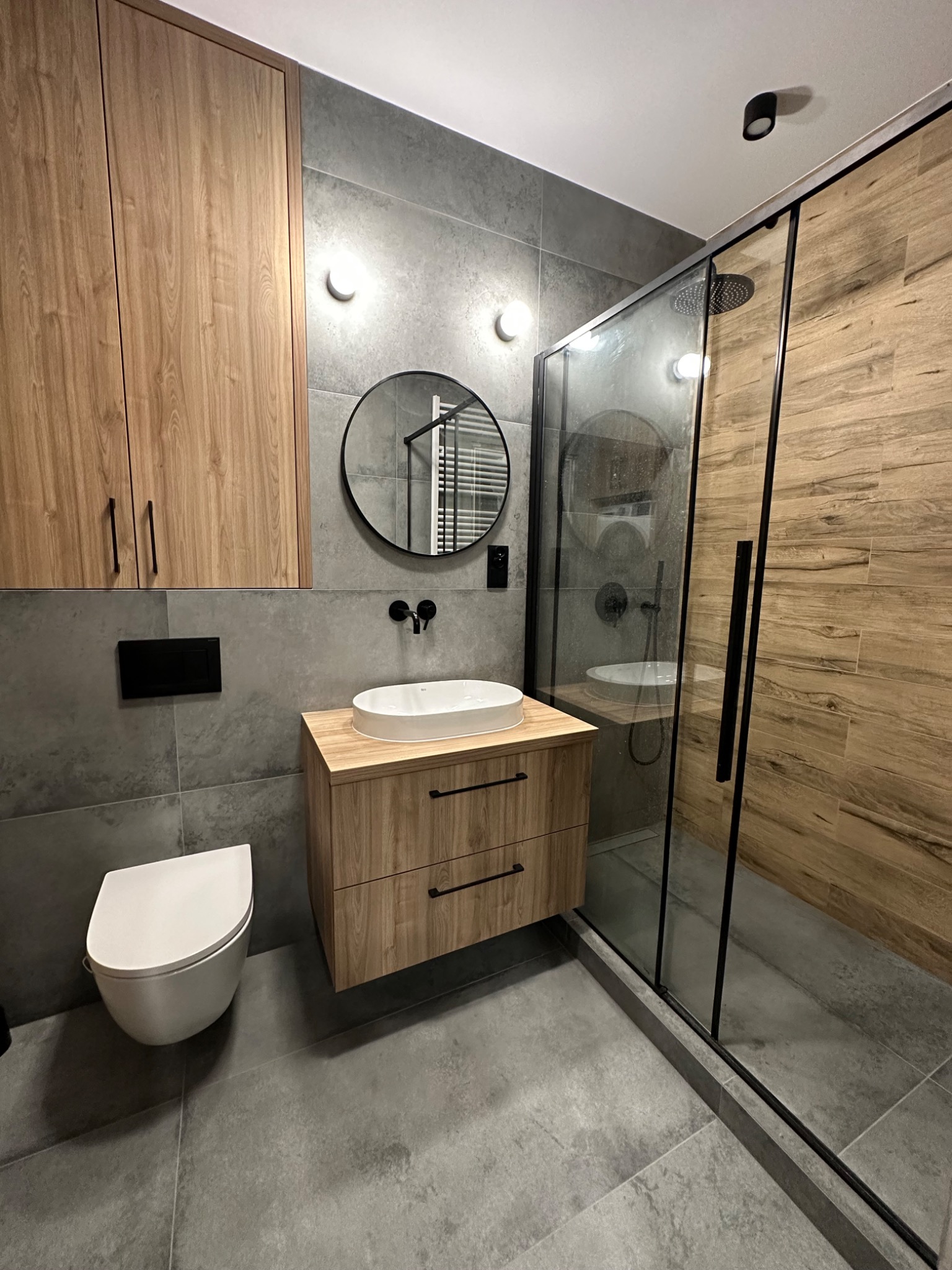Nowoczesna łazienka z szarymi płytkami, drewnianymi szafkami i kabiną prysznicową typu walk-in z elementami drewna, okrągłe lustro nad umywalką, podwieszana toaleta.