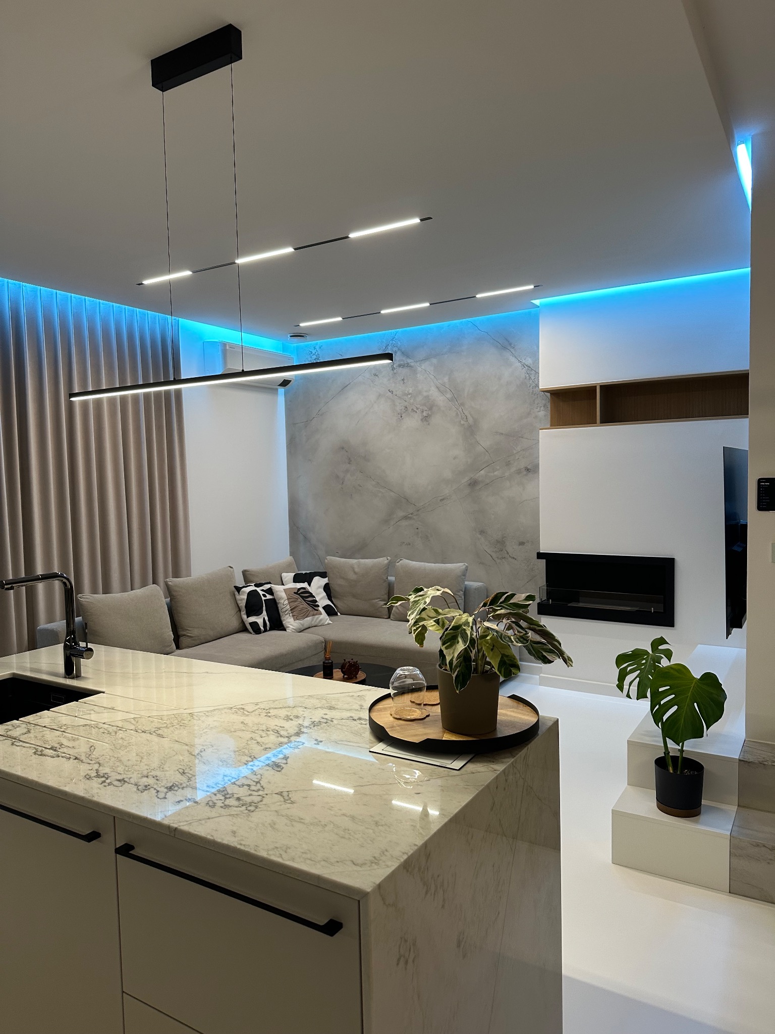 Nowoczesne wnętrze: marmurowa ściana, podświetlenie LED, wyspa kuchenna z blatem imitującym marmur, szara sofa i minimalistyczny kominek. Rośliny doniczkowe dodają życia.