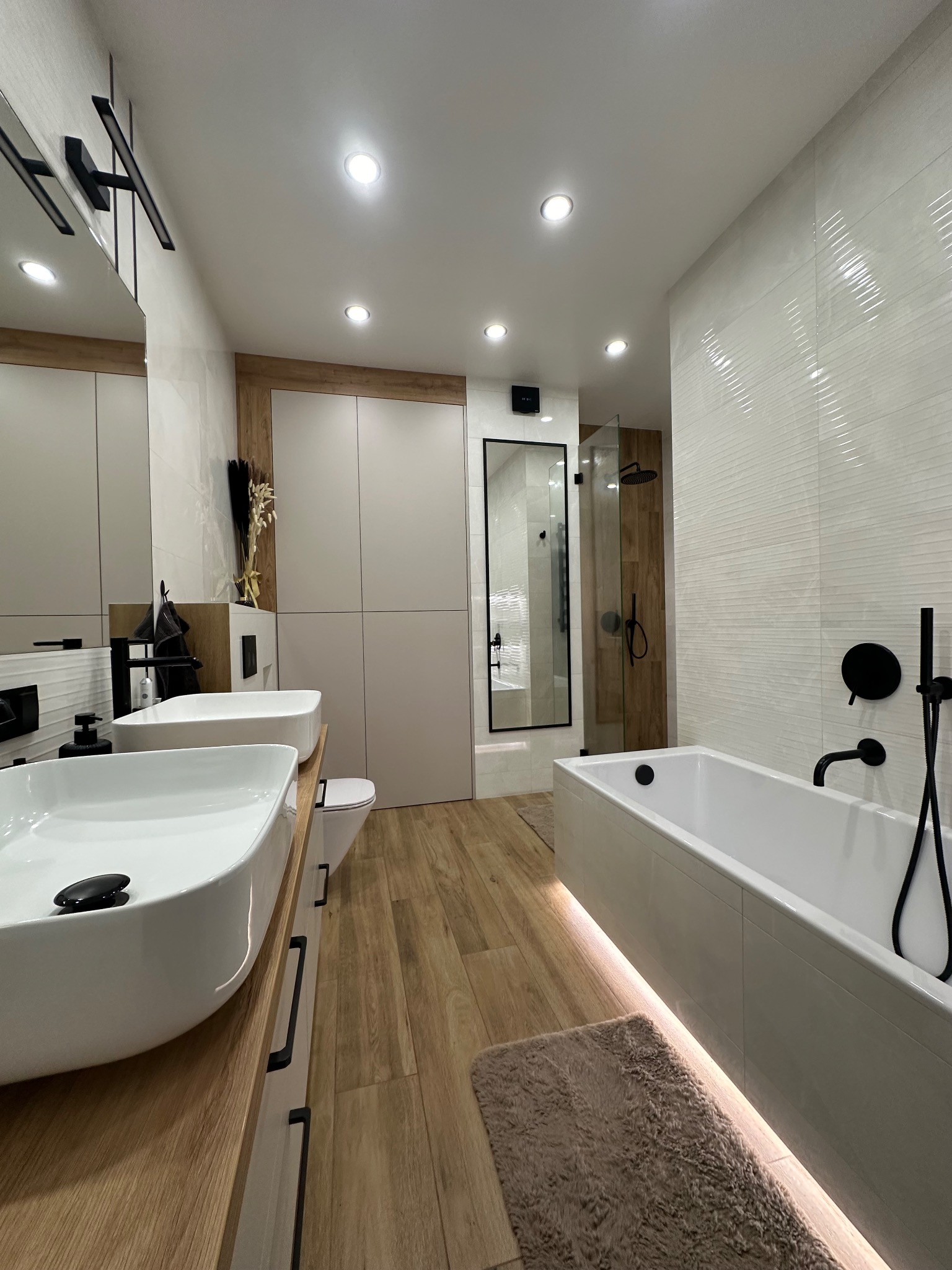 Nowoczesna łazienka z drewnianymi akcentami: podwójna umywalka nablatowa, wanna z podświetleniem LED, kabina prysznicowa w czarnej ramie i płytki imitujące drewno.