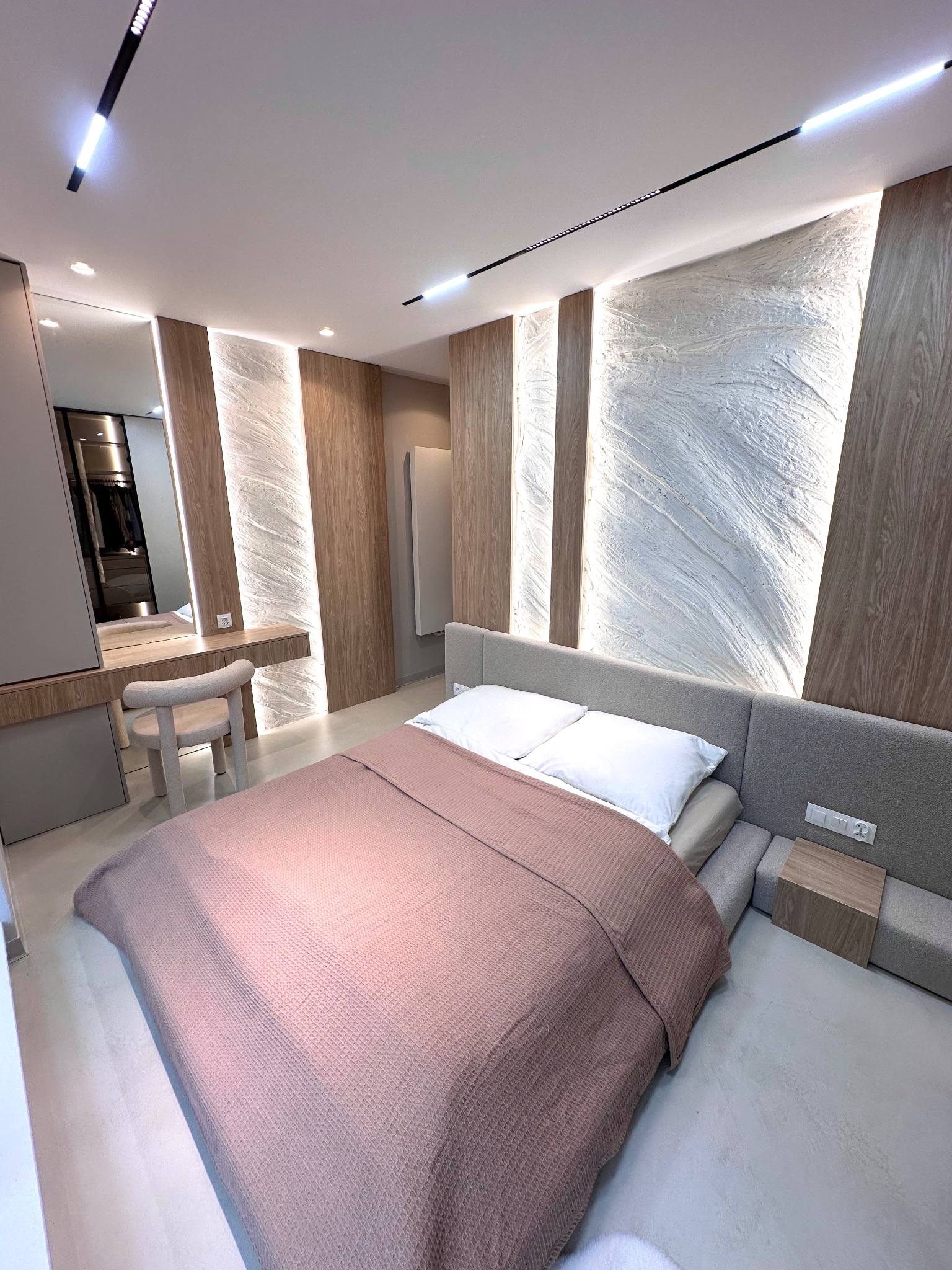 Nowoczesna sypialnia z łóżkiem, toaletką i oświetleniem LED. Ściana z dekoracyjną strukturą i drewnianymi panelami. Minimalistyczny design, jasne kolory, eleganckie wykończenie.