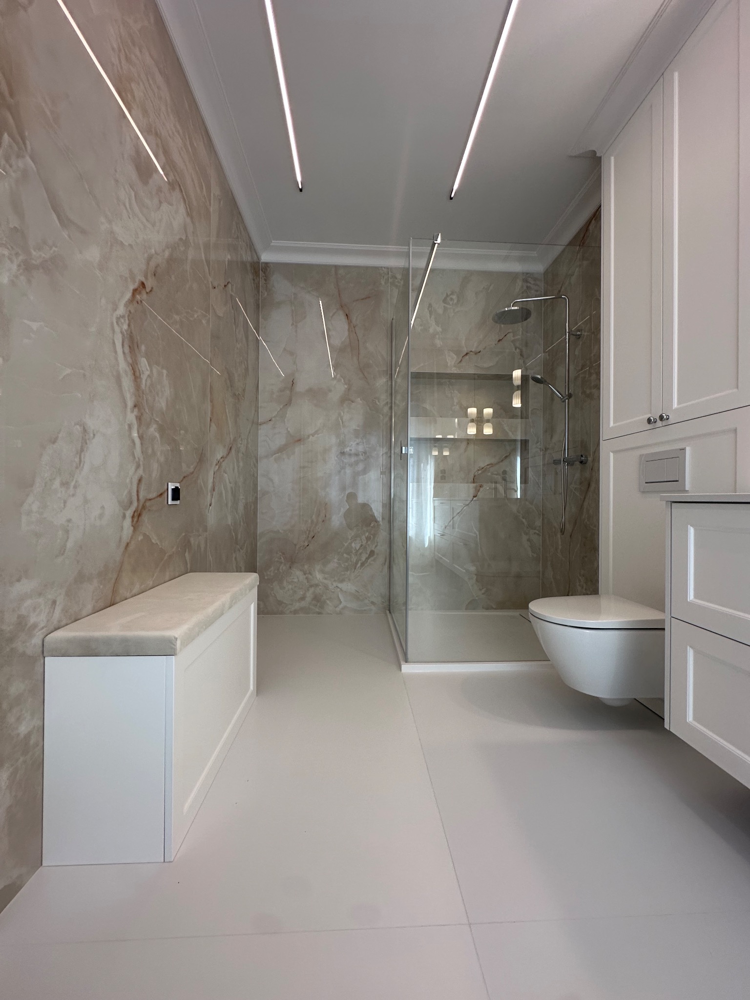 Elegancka łazienka z marmurowymi płytkami, kabiną prysznicową typu walk-in, podświetleniem LED i białą zabudową meblową z siedziskiem. Minimalistyczny design.