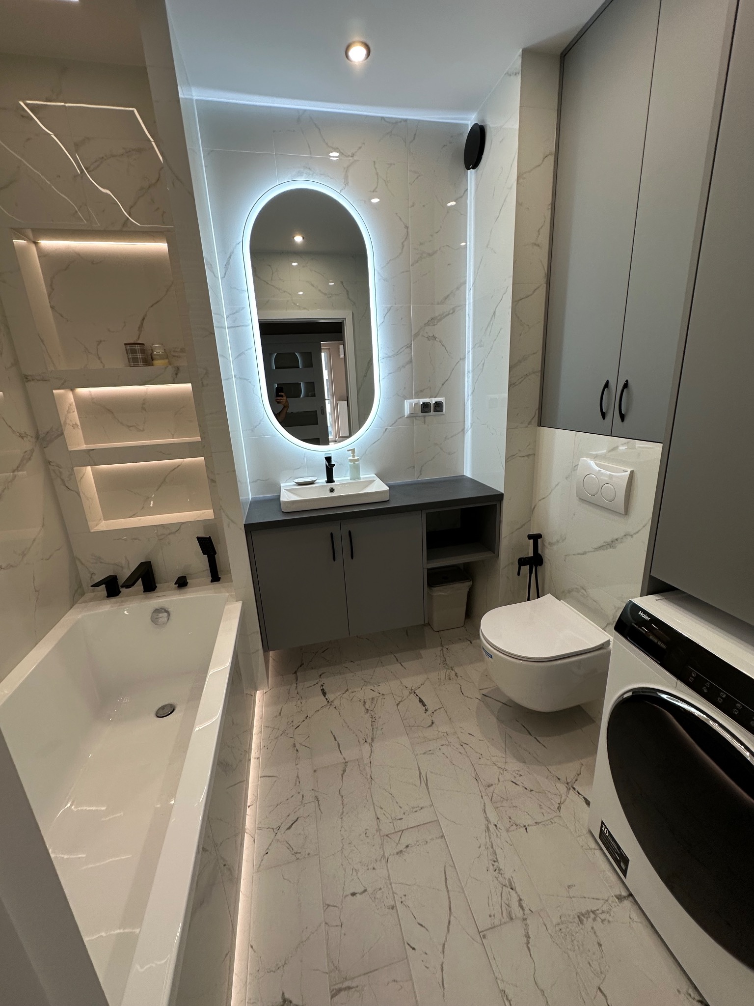 Nowoczesna łazienka z wanną, umywalką z podświetlanym lustrem, toaletą i pralką. Białe płytki imitujące marmur, szare szafki i czarne akcenty.