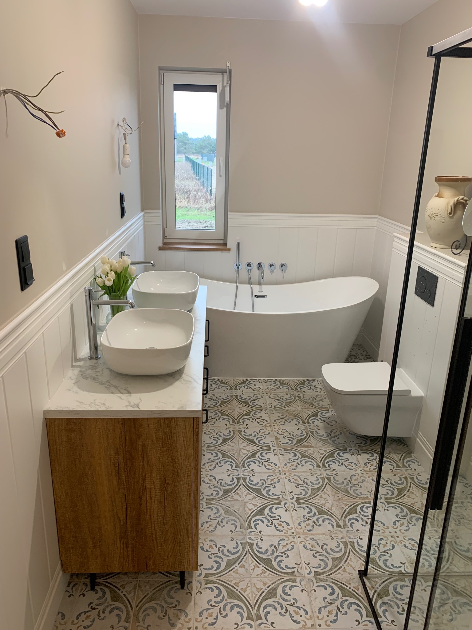 Jasna łazienka z wolnostojącą wanną, dwoma umywalkami na drewnianej szafce z marmurowym blatem i wzorzystą podłogą. Widoczne okno i fragment lustra.