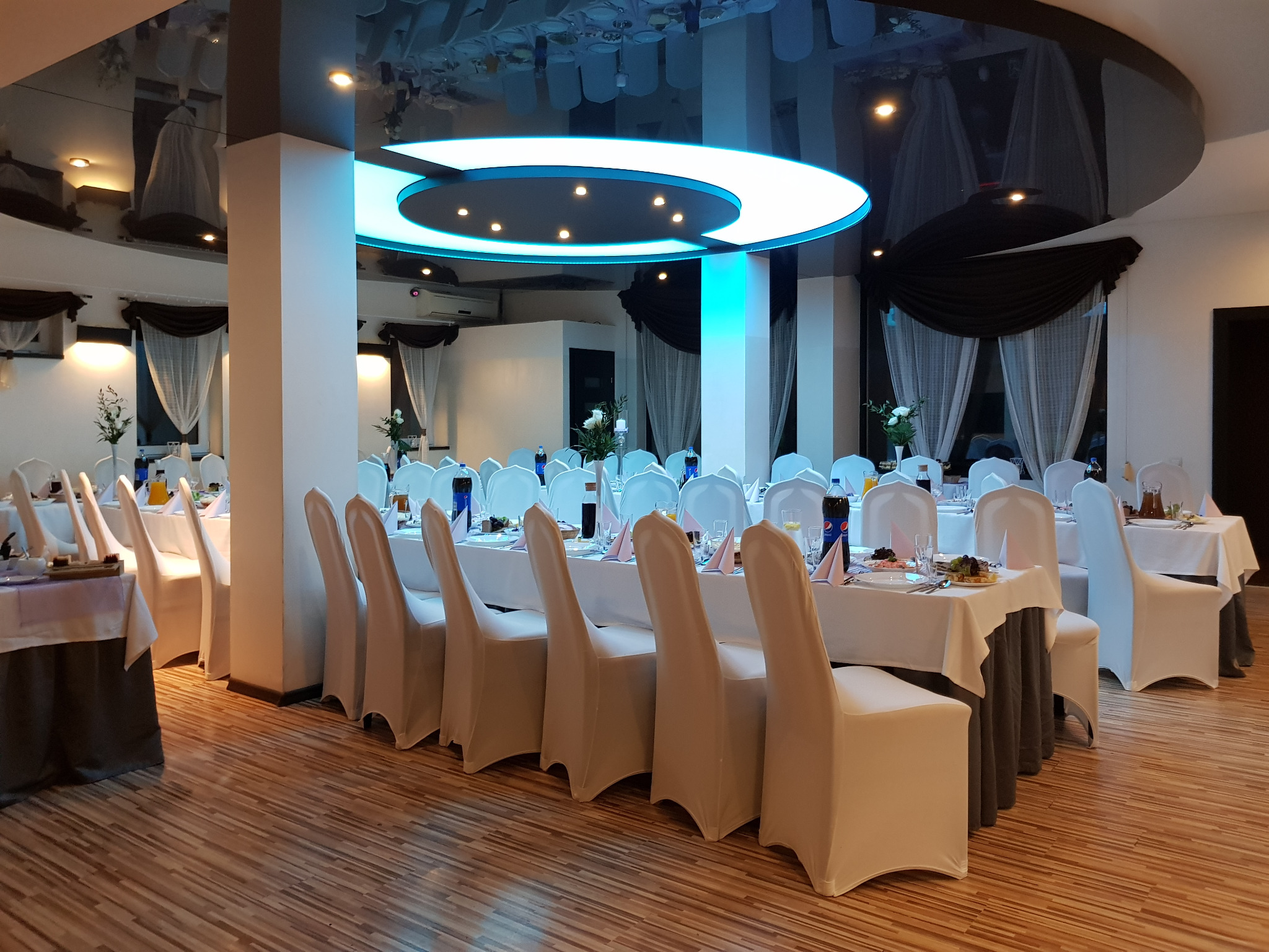 Sala bankietowa z nakrytymi stołami i krzesłami w białych pokrowcach, oświetlona nowoczesną lampą sufitową w kształcie okręgu.