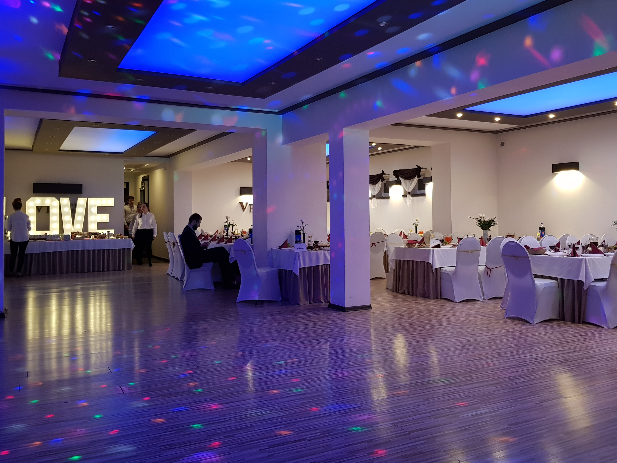 Sala bankietowa z dekoracją świetlną, stołami z białymi obrusami i krzesłami w pokrowcach, napis LOVE podświetlony z tyłu.