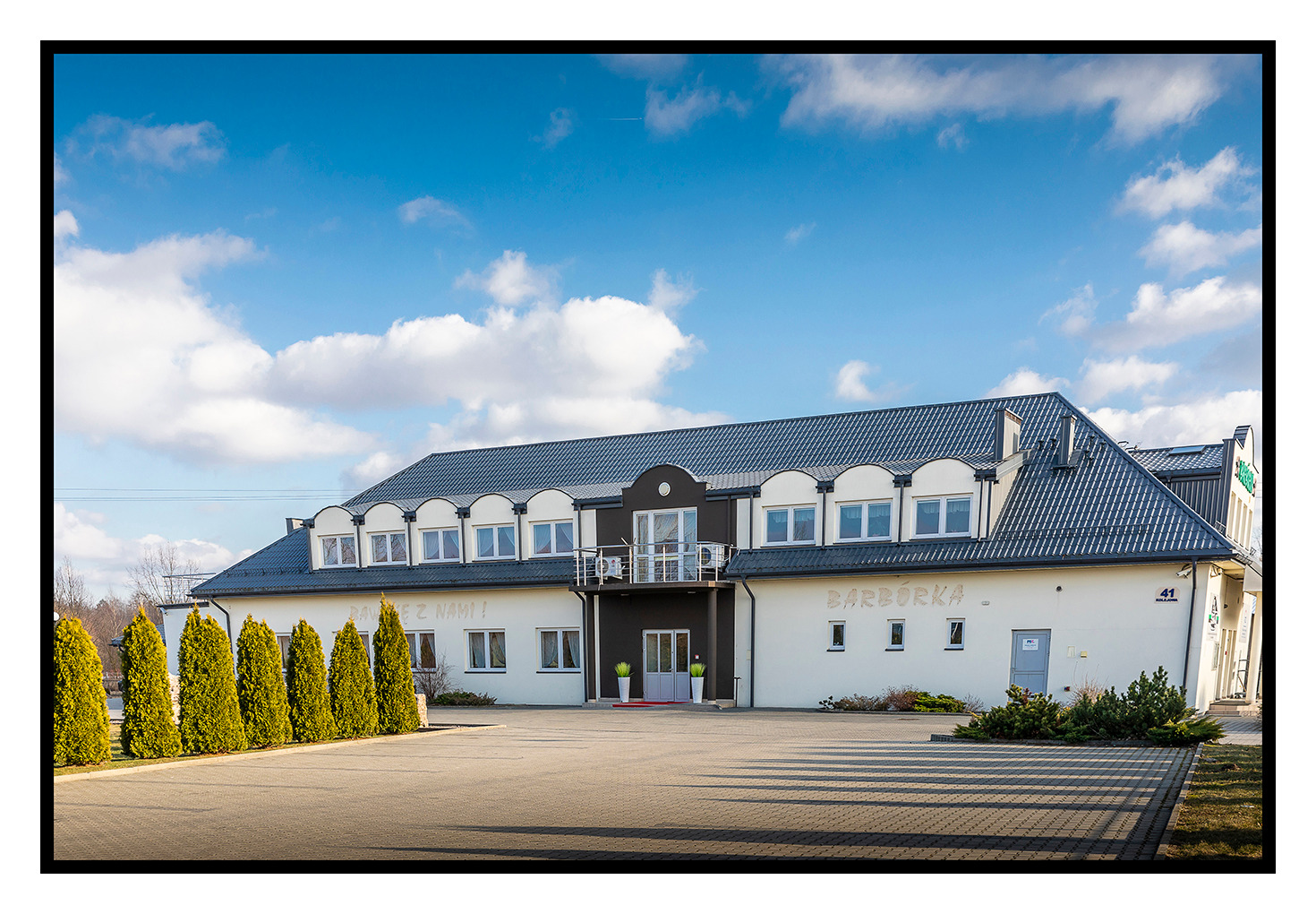 Dwupiętrowy budynek z szarym dachem, białymi ścianami i balkonem, przed budynkiem rząd zielonych iglaków i wybrukowany plac, na ścianie napis 'Barbórka'.