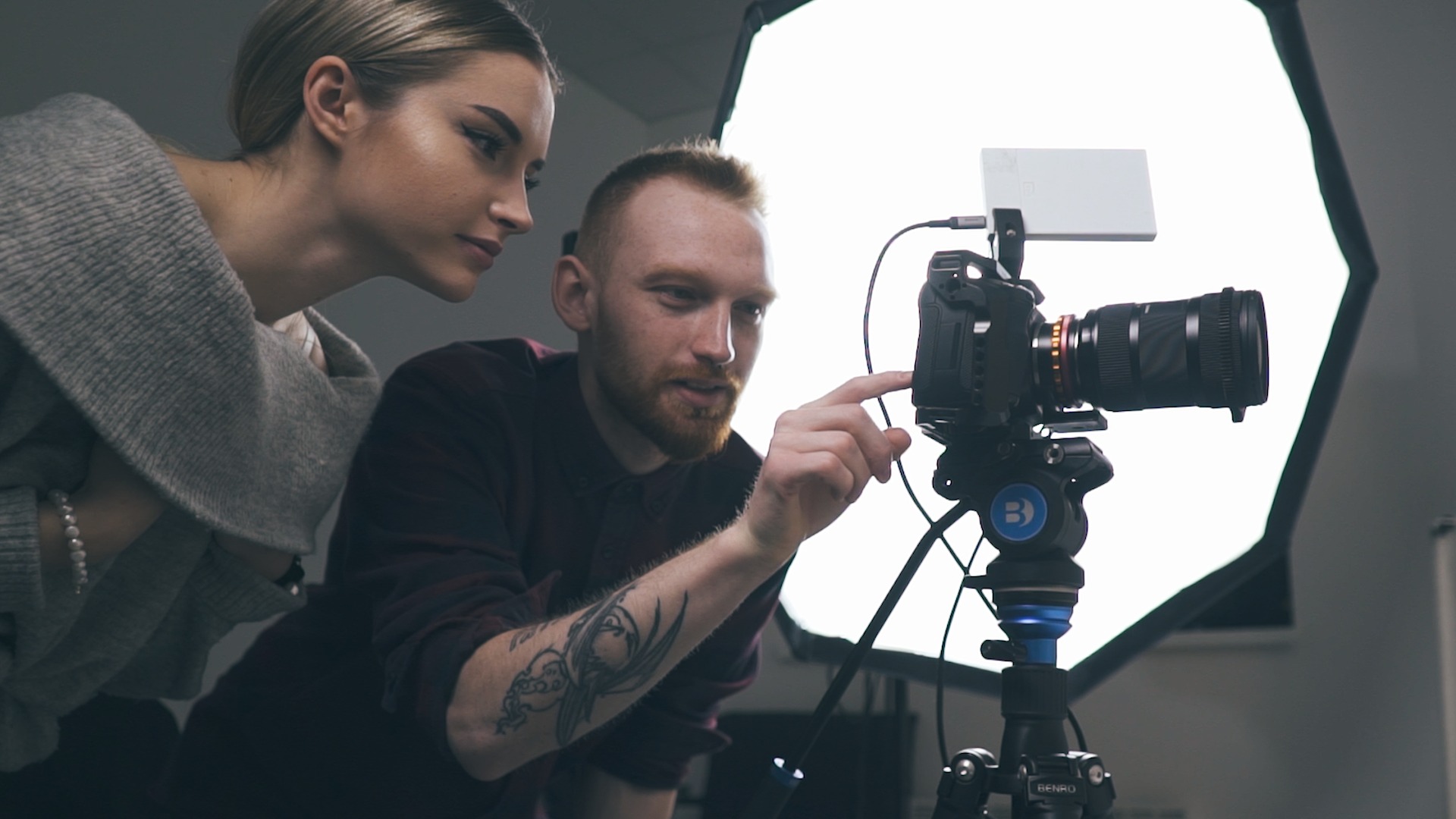 Operator z tatuażem na ramieniu i asystentka sprawdzają ustawienia kamery na statywie przed softboxem.