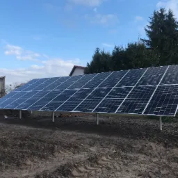 Instalacja paneli fotowoltaicznych na metalowych podporach na gruncie z widocznym pracownikiem w tle, dom i drzewa w tle, pochmurne niebo.