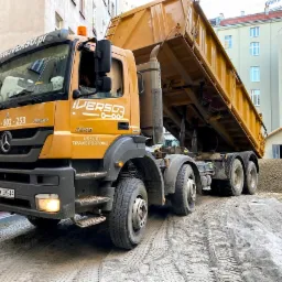 Żółta wywrotka Mercedes-Benz z podniesioną skrzynią ładunkową, wysypująca żwir na plac budowy w otoczeniu budynków mieszkalnych.