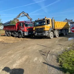 Ciężarówka z wywrotką i koparka załadowują urobek w ziemi we Wrocławiu, widoczne dwie ciężarówki z logo firmy.