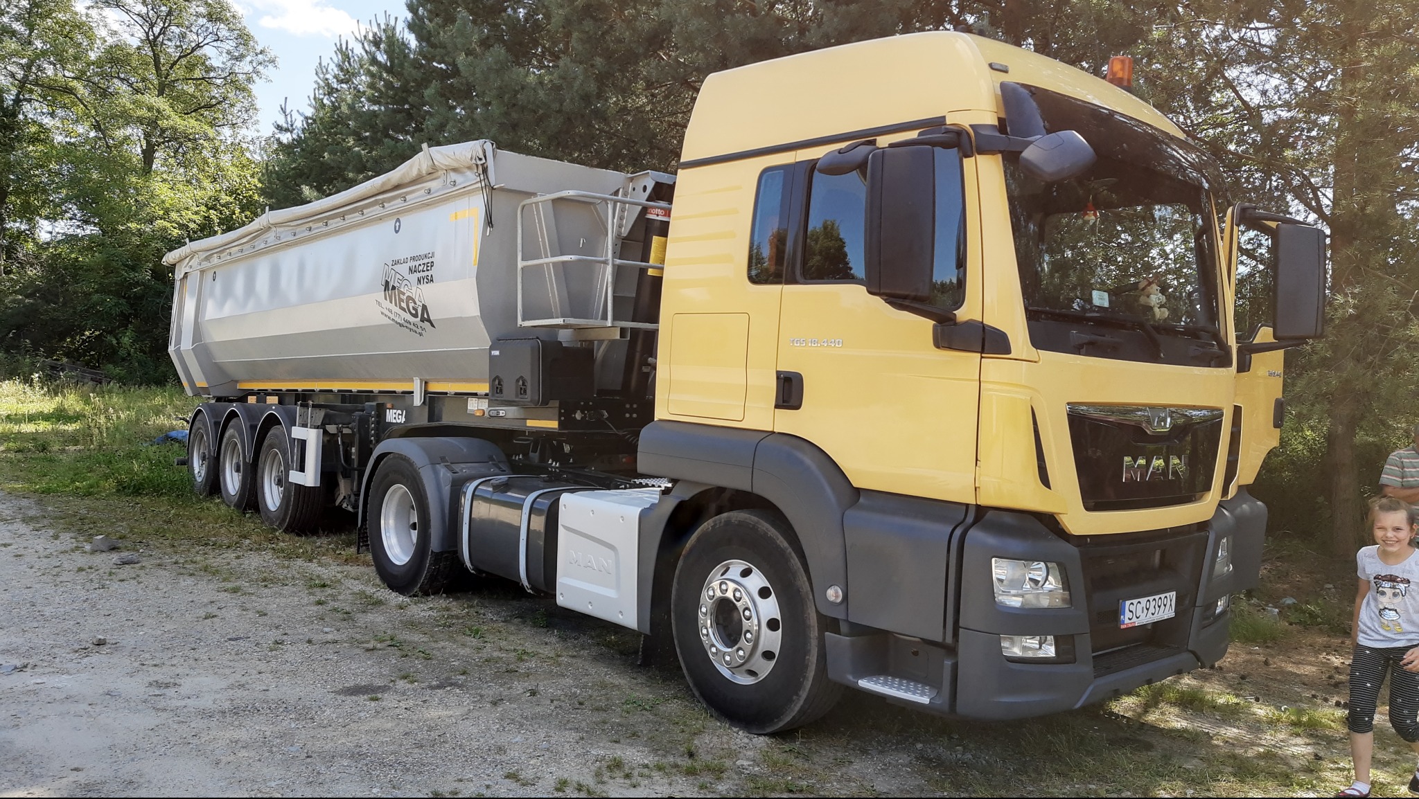 Żółta ciężarówka MAN z naczepą MEGA na szutrowej drodze, widok z boku, na pierwszym planie dziewczynka.