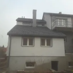 Dwukondygnacyjny budynek z szarą dachówką i jasną elewacją, widoczne okna, balkon z czarną balustradą i komin, na dole widoczny fragment fundamentu i wejście do piwnicy.