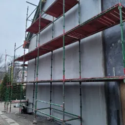 Budynek w trakcie prac elewacyjnych, widoczne rusztowania, ocieplenie na fragmencie ściany i świeżo nałożony tynk na pozostałej części.