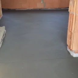 Świeżo wylana, gładka, szara posadzka w surowym wnętrzu budynku z czerwonej cegły, widoczne fragmenty ścian i progi drzwiowe.