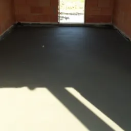 Świeżo wylana, gładka posadzka w surowym stanie, z widocznymi ceglanymi ścianami i wpadającym światłem słonecznym przez otwór drzwiowy, ukazującym teren zewnętrzny.