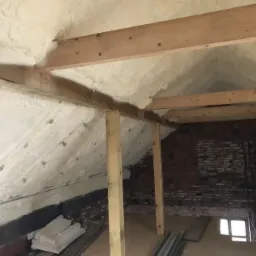 Poddasze z drewnianą konstrukcją krokwiową, pokryte białą, grubą warstwą izolacji natryskowej z pianki PUR, widoczne ślady po dyfuzorach, w tle fragment ściany z czerwonej cegły.