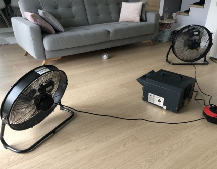 Ozonowanie mieszkania: widok na szary salon z dwoma wentylatorami podłogowymi i generatorem ozonu na drewnianej podłodze.