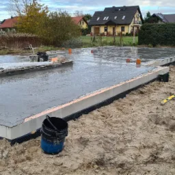 Wylany fundament z izolacją przeciwwilgociową, widoczne wystające pręty zbrojeniowe i narzędzia budowlane na placu budowy domu jednorodzinnego.