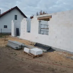 Wznoszenie ścian domu z białych bloczków, widoczne otwory okienne i drzwiowe, w tle istniejący dom z ciemnym dachem, na pierwszym planie paleta z bloczkami i ułożone betonowe nadproża.