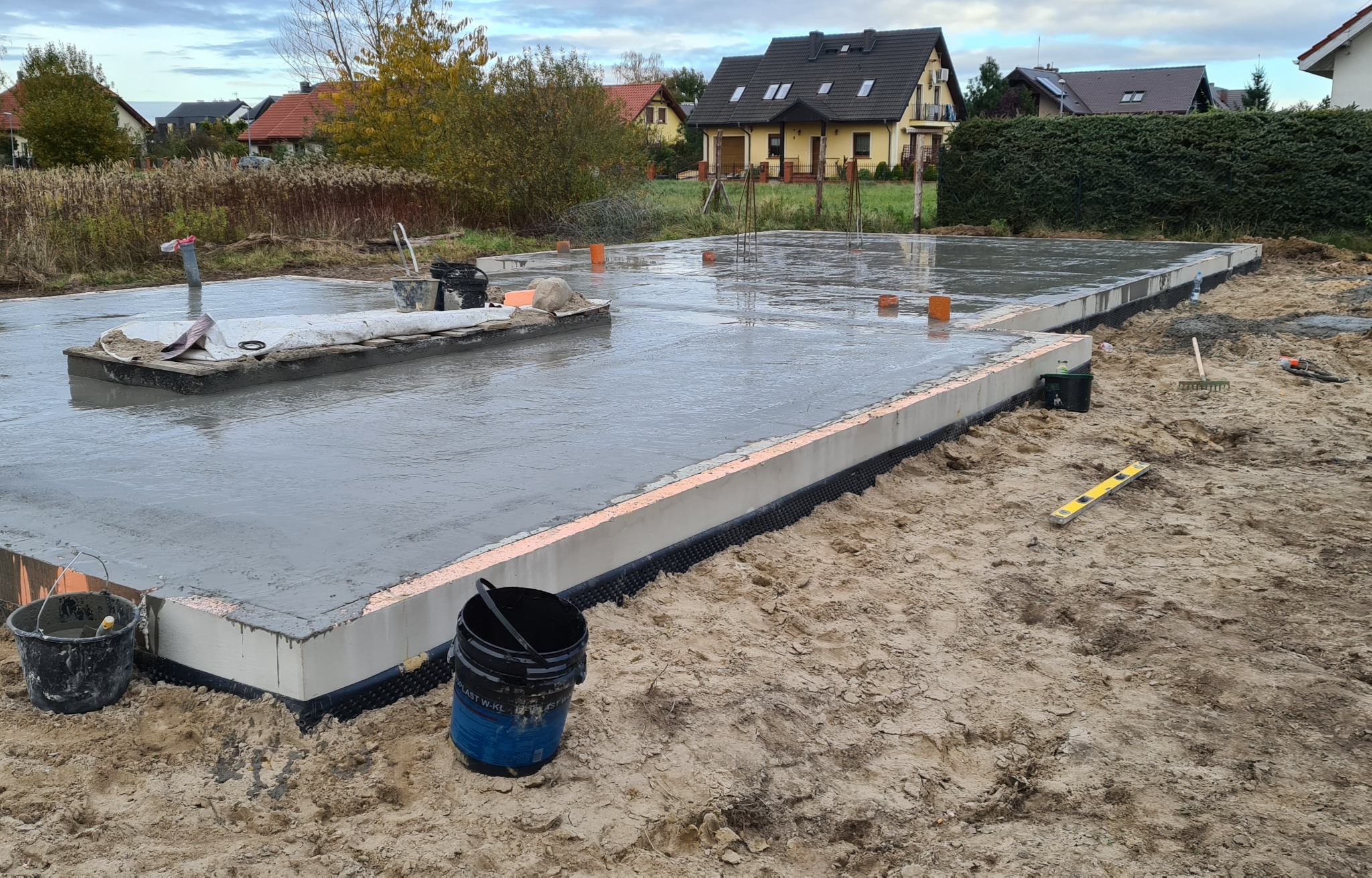 Wylany fundament z izolacją przeciwwilgociową, widoczne wystające pręty zbrojeniowe i narzędzia budowlane na placu budowy domu jednorodzinnego.