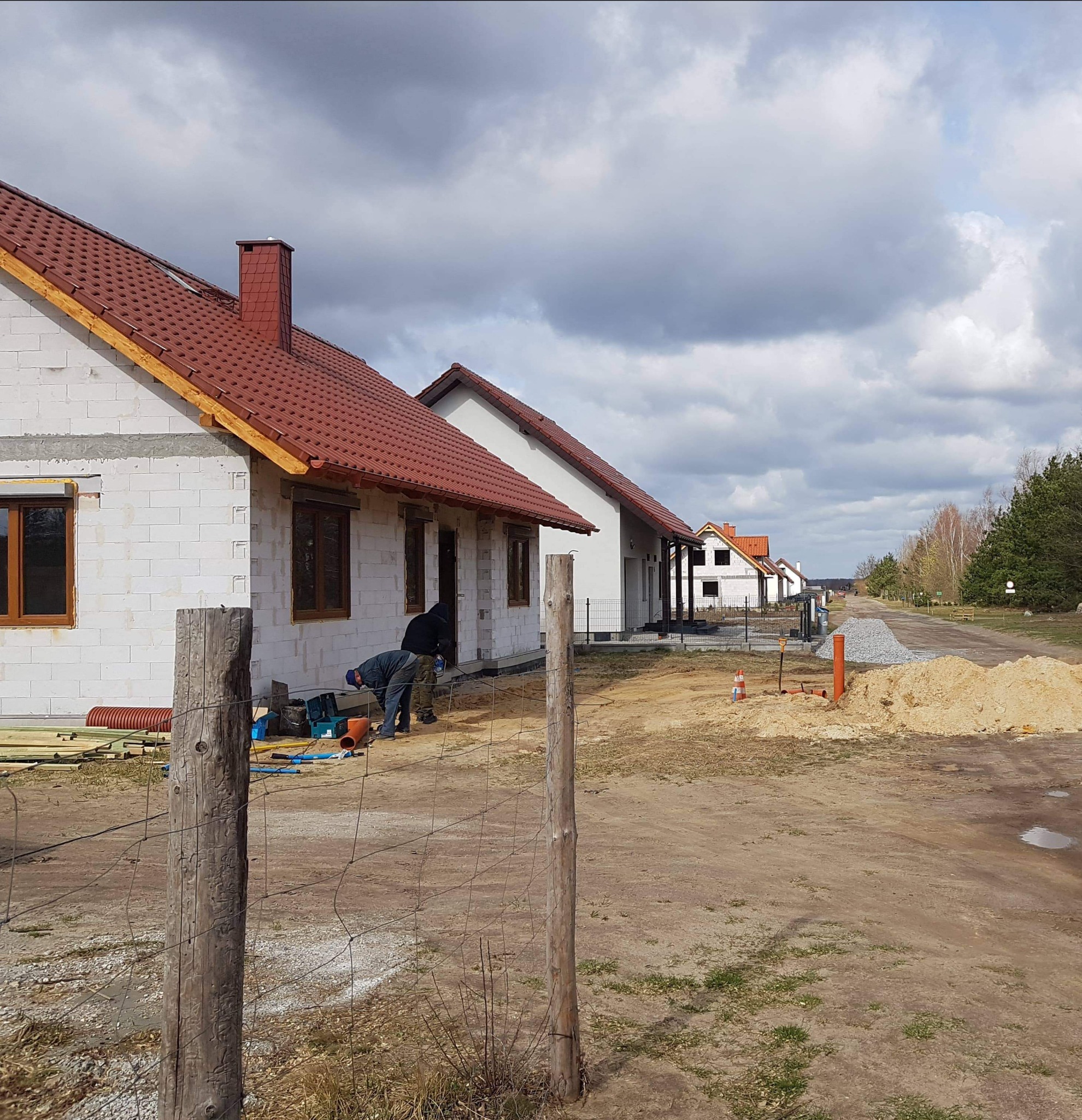 Budowa osiedla domów jednorodzinnych z białego bloczka, widoczni dwaj robotnicy pracujący przy instalacjach zewnętrznych, w tle kolejne domy w różnym stopniu zaawansowania.