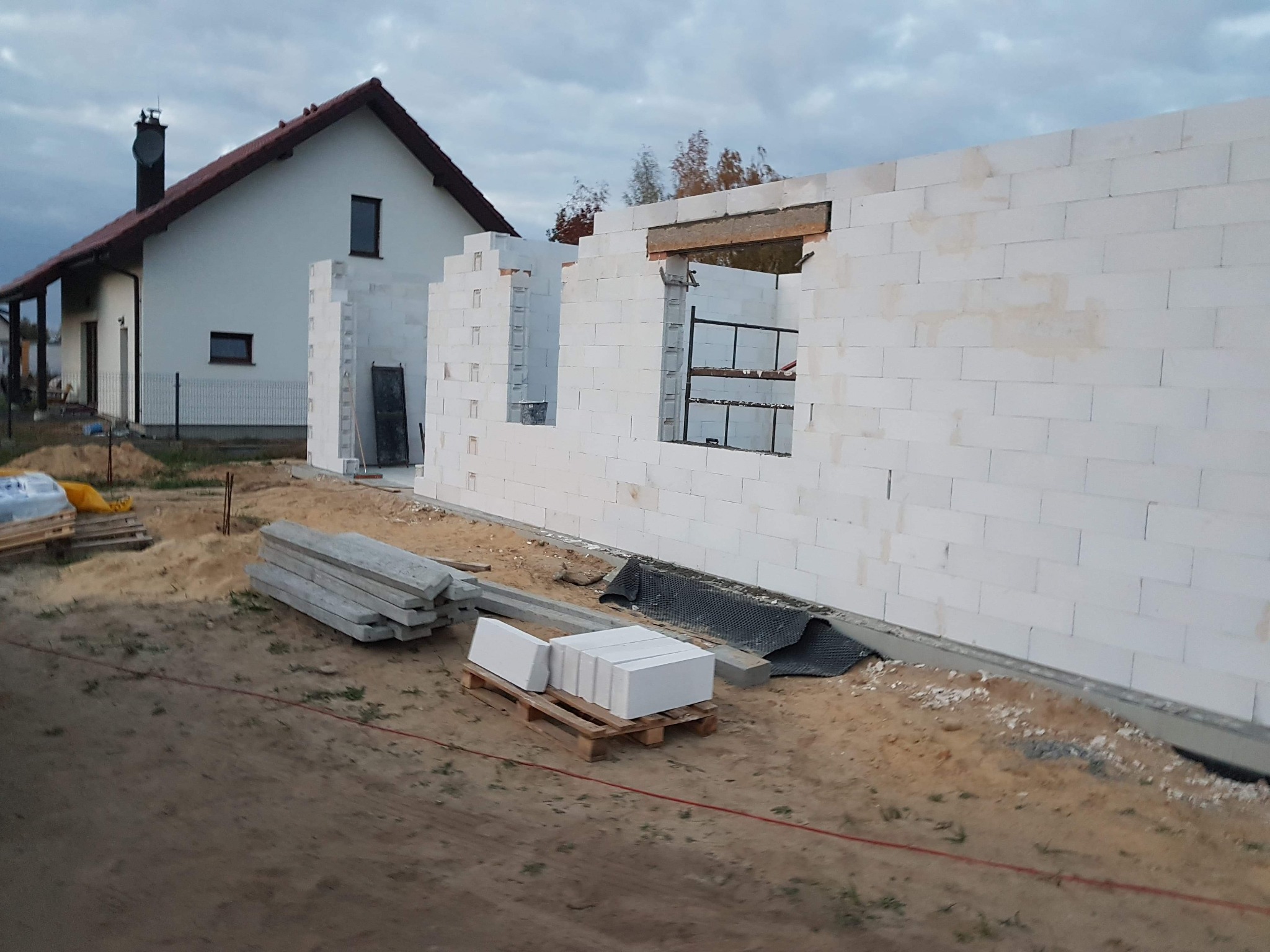 Wznoszenie ścian domu z białych bloczków, widoczne otwory okienne i drzwiowe, w tle istniejący dom z ciemnym dachem, na pierwszym planie paleta z bloczkami i ułożone betonowe nadproża.