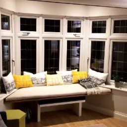 Wnęka okienna z siedziskiem i poduszkami w odcieniach żółci i szarości, jasna podłoga i dywan, widok na wnętrze domu.