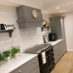 Szara kuchnia na wymiar z czarnym piecem kaflowym, białymi płytkami metra i marmurowym blatem, uzupełniona o zioła w doniczkach i dekoracje na półkach.