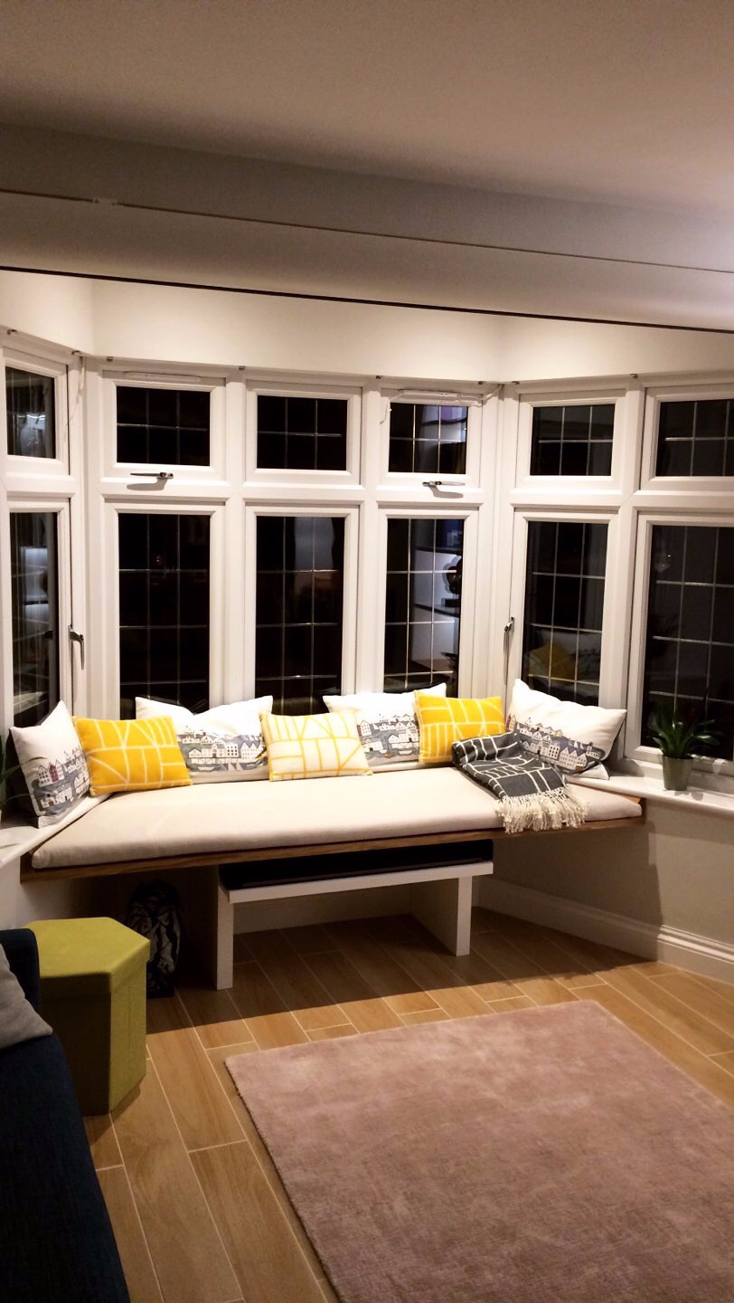 Wnęka okienna z siedziskiem i poduszkami w odcieniach żółci i szarości, jasna podłoga i dywan, widok na wnętrze domu.